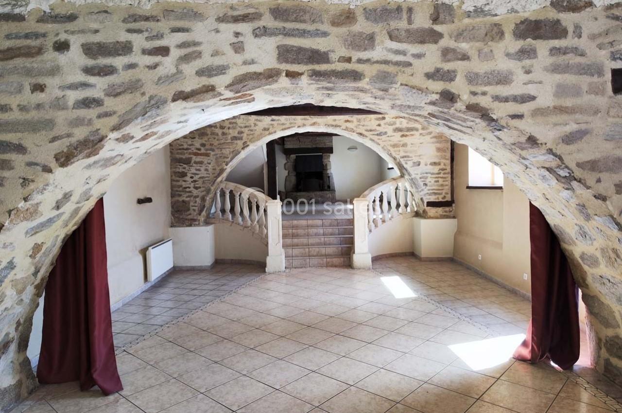 Salle voûtée en pierre avec sol carrelé, escaliers menant à une cheminée et rideaux rouges encadrant les ouvertures.