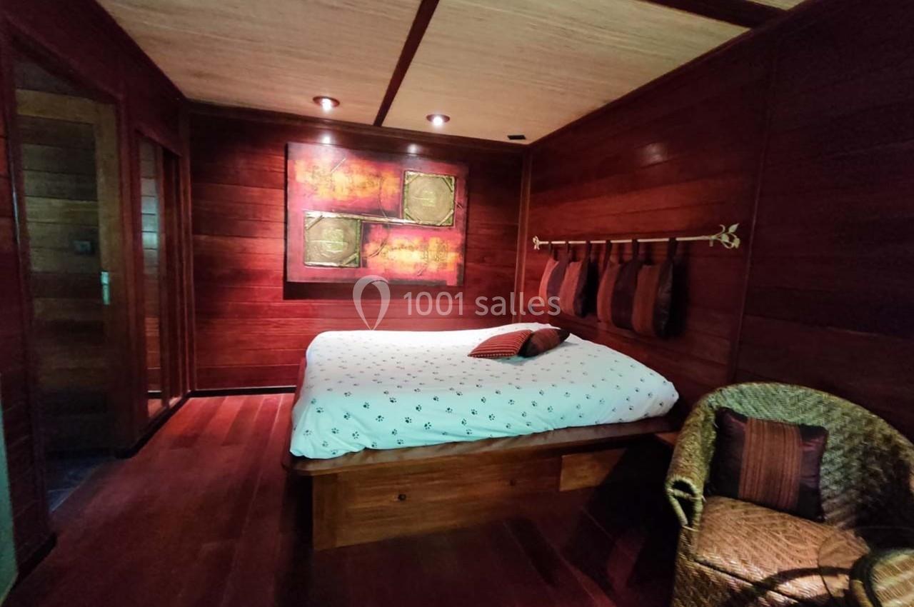 Chambre en bois avec lit double, coussins décoratifs, fauteuil en osier et tableau coloré au mur.