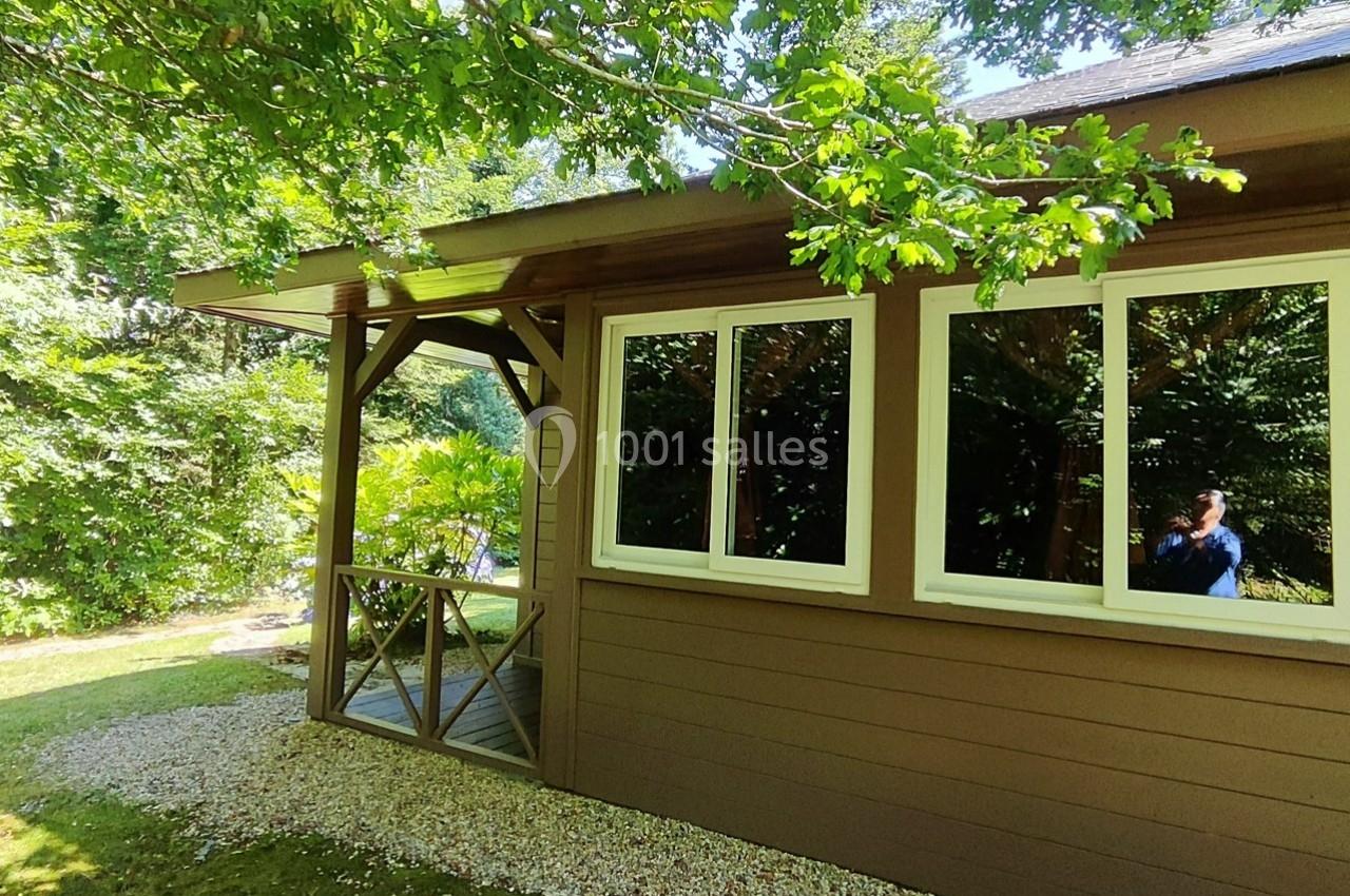 Maison en bois avec terrasse couverte, entourée de verdure et éclairée par la lumière naturelle.