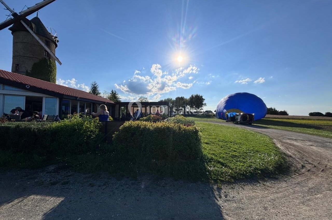 Un moulin près d'une terrasse ensoleillée, avec une montgolfière bleue en préparation sur un terrain herbeux.