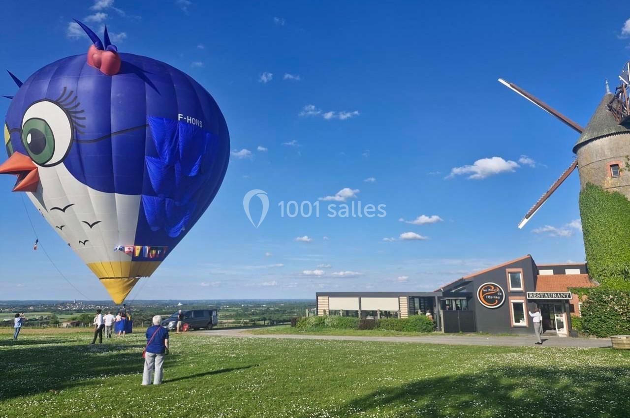 Montgolfière colorée en forme d'oiseau sur un terrain herbeux près d'un moulin et d'un bâtiment moderne par temps clair.