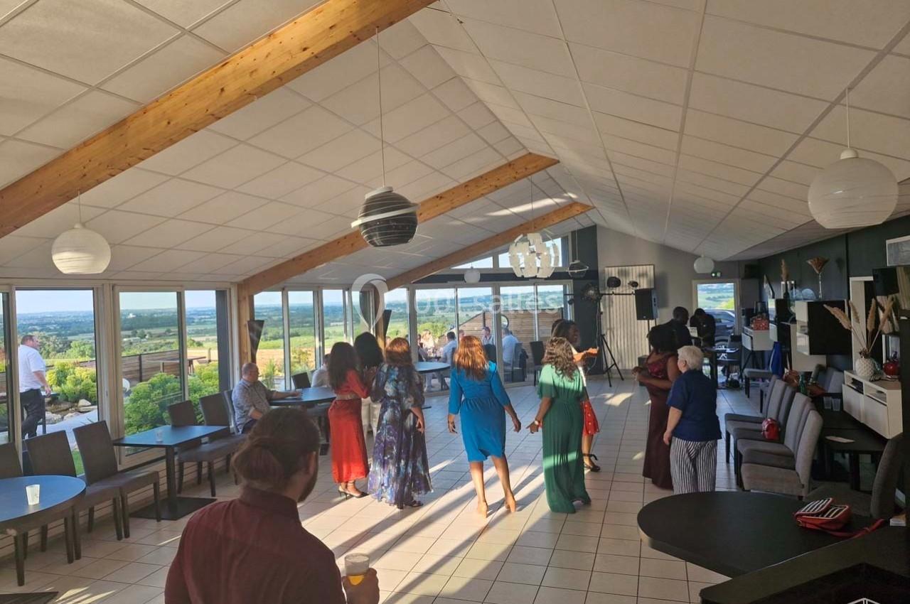 Des personnes discutent et dansent dans une salle lumineuse avec de grandes baies vitrées offrant une vue sur la campagne.