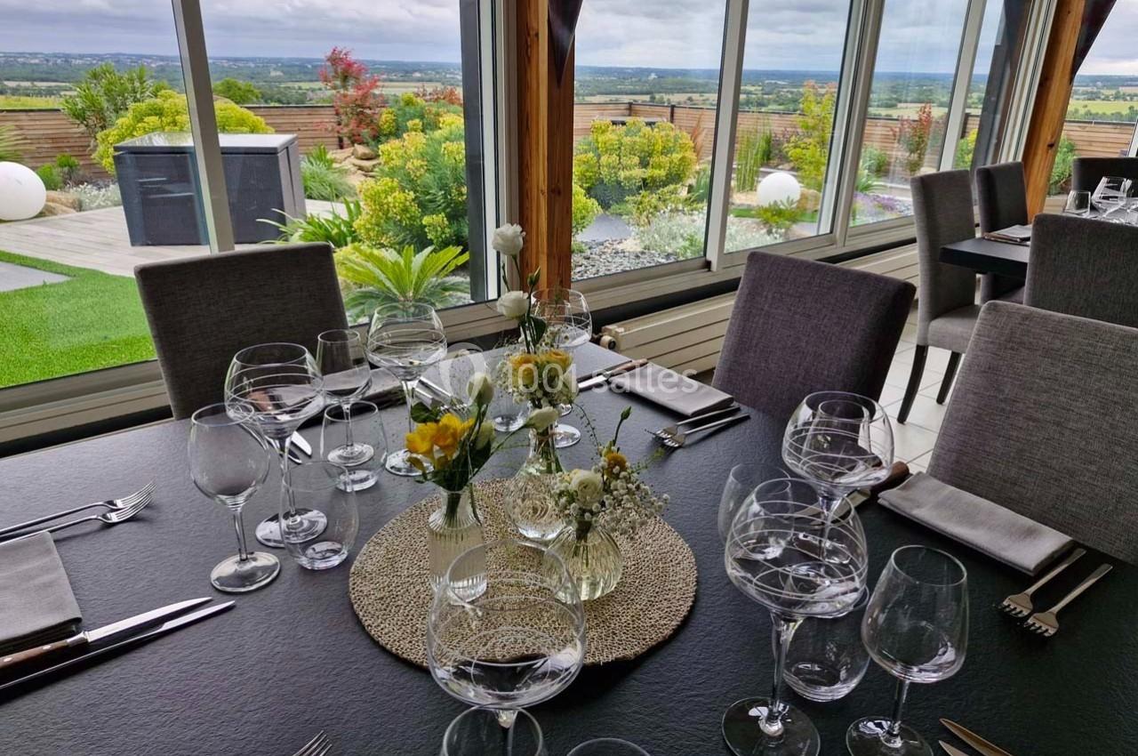 Table élégamment dressée avec des verres et des fleurs, donnant sur un jardin et une vue panoramique à travers de grandes…