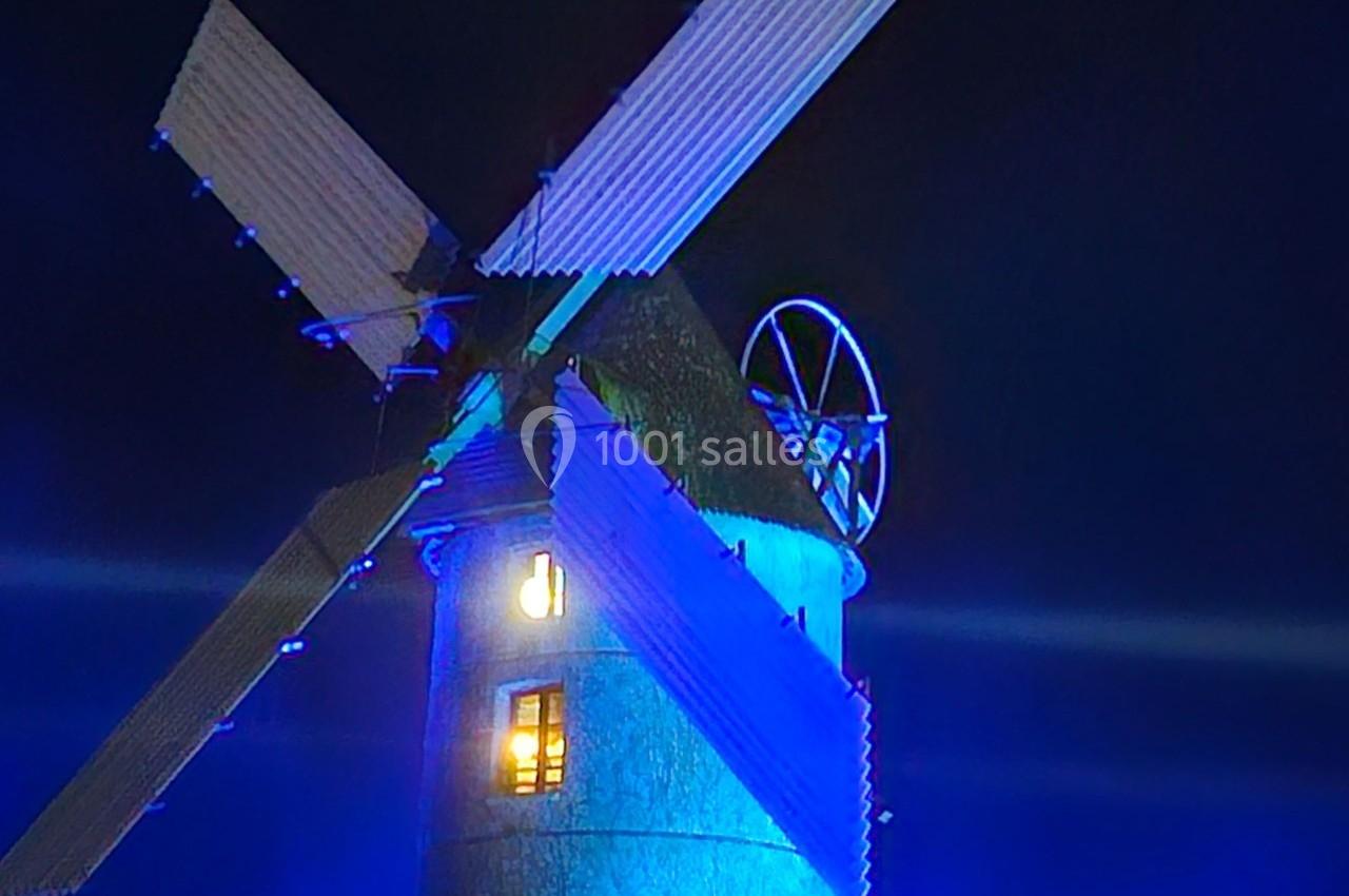 Moulin éclairé en bleu avec des pales blanches, visible de nuit, une fenêtre illuminée au centre.