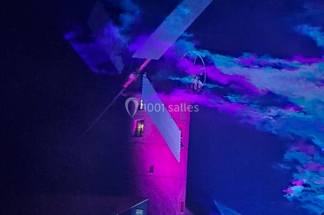 Moulin éclairé par des lumières violettes et bleues, avec des effets de fumée colorée dans un décor nocturne.