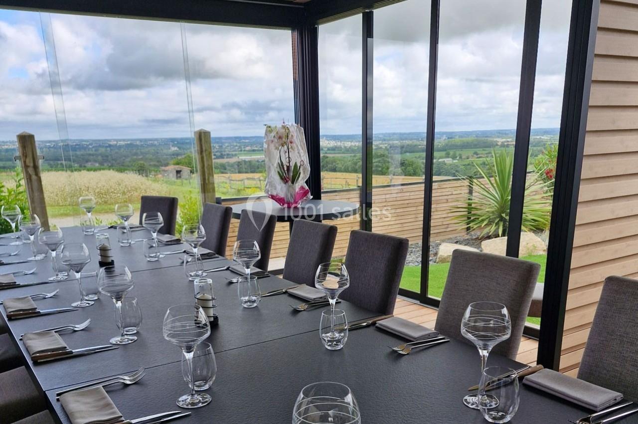 Salle à manger avec grande table dressée, baies vitrées offrant une vue panoramique sur la campagne verdoyante.