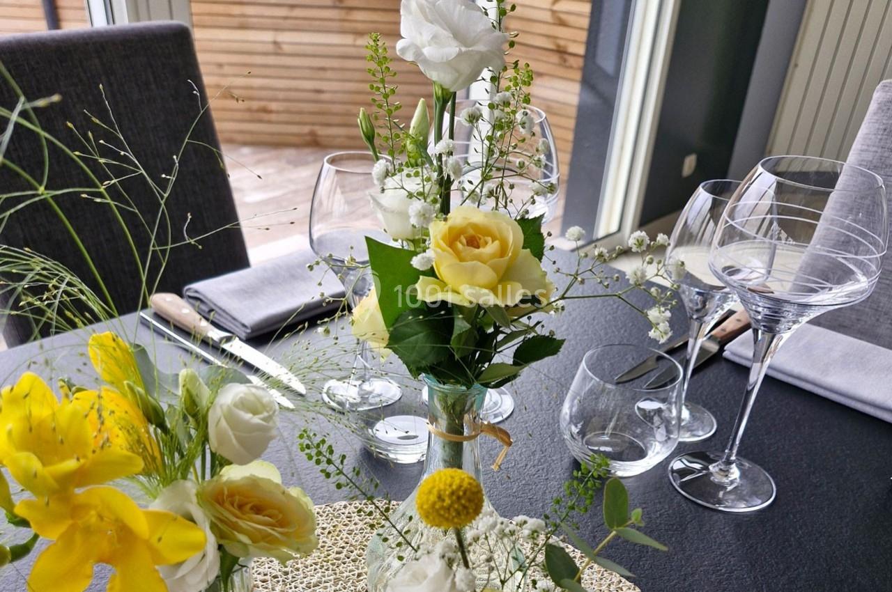 Table dressée avec des fleurs jaunes et blanches, verres à vin et serviettes grises, près d'une terrasse en bois.