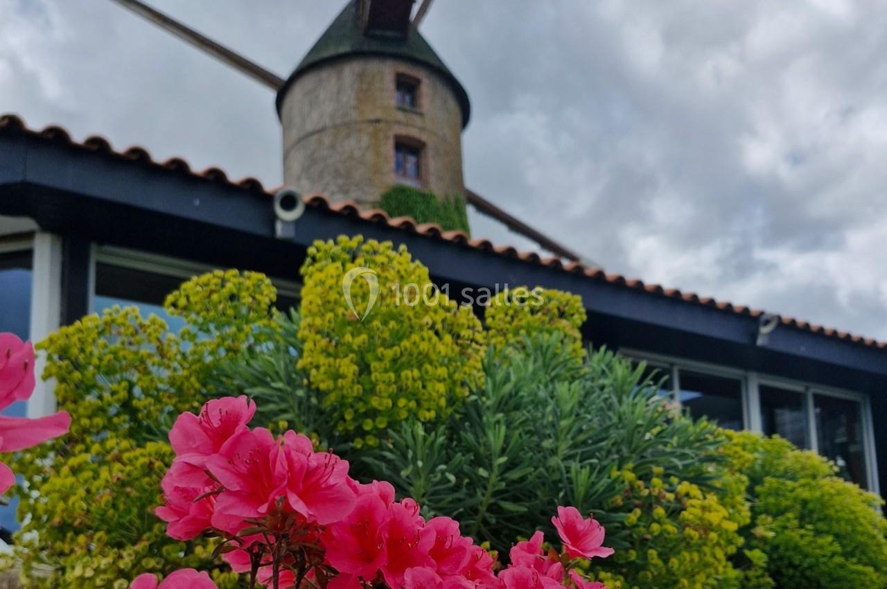 Fleurs roses au premier plan devant un moulin en pierre avec des ailes en bois, sous un ciel nuageux.