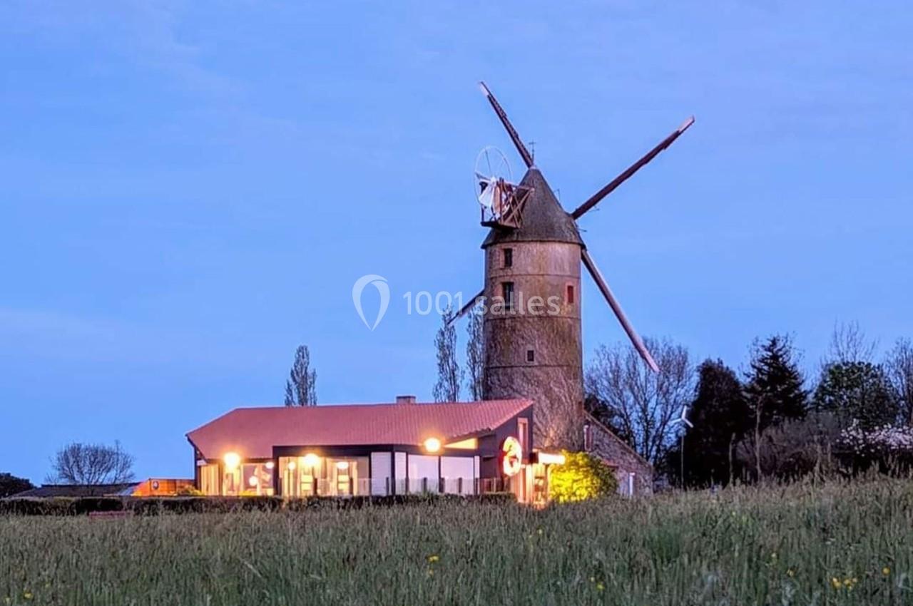 Un moulin à vent en pierre près d'une maison éclairée, entouré de champs et d'arbres au crépuscule.