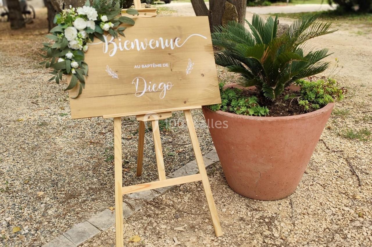 Panneau en bois sur chevalet avec fleurs, inscrit ’Bienvenue au baptême de Diego’, placé dans un jardin.