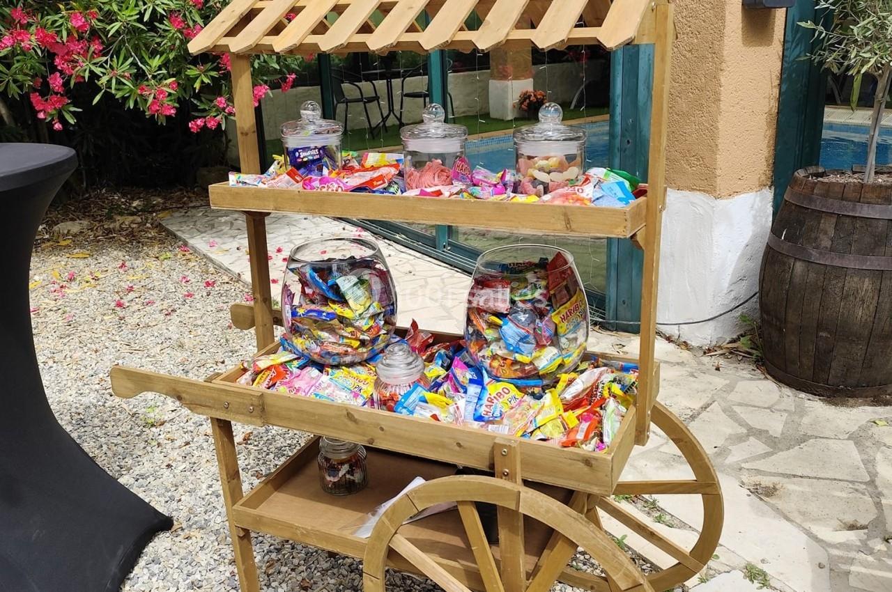 Chariot en bois rempli de bonbons colorés dans des bocaux et sachets, placé sur un sol en gravier près d'une végétation…