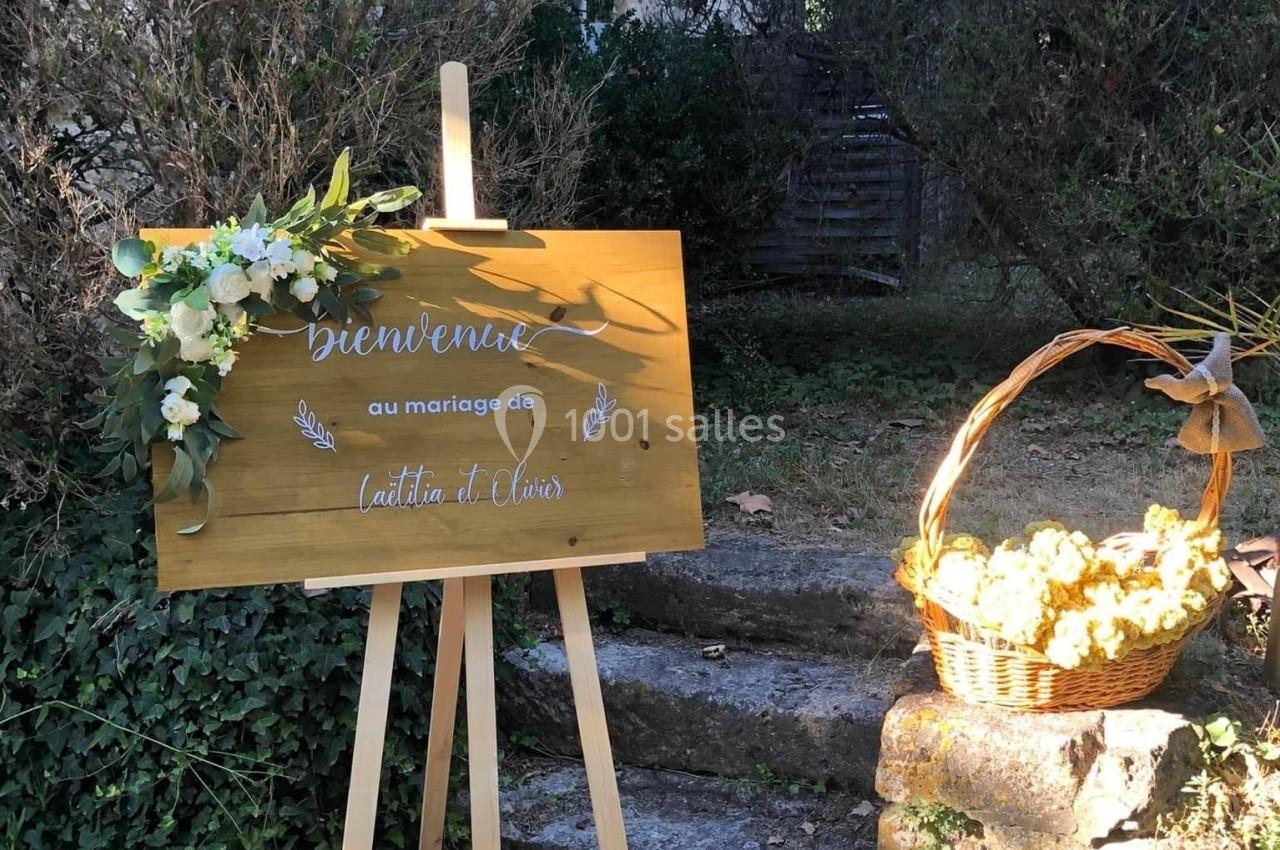 Panneau en bois décoré de fleurs blanches, posé sur un chevalet, à côté d'un panier de fleurs jaunes dans un jardin.