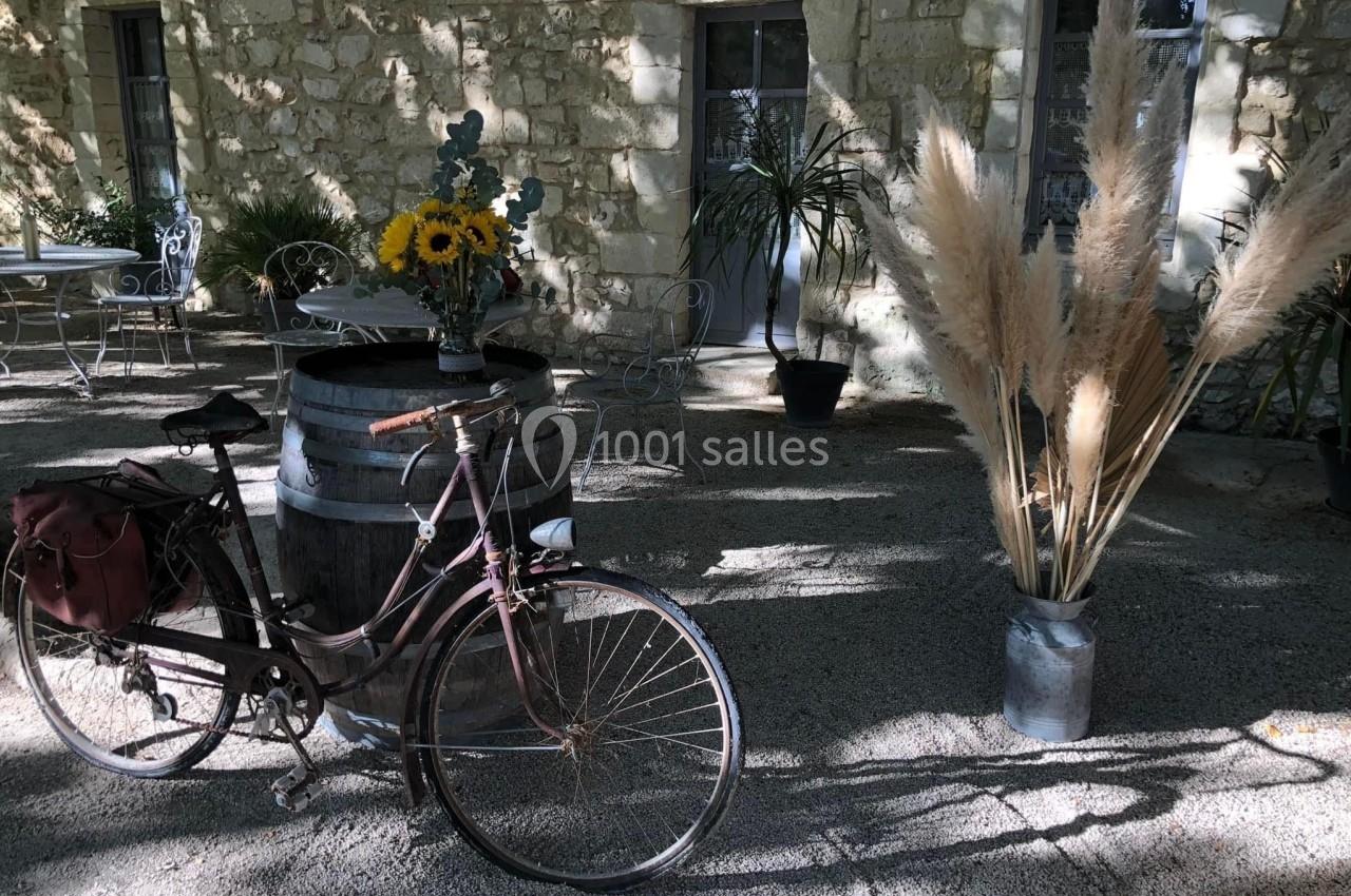 Un vélo ancien posé près d'un tonneau décoré de fleurs, devant un mur en pierre et des plantes en pot.