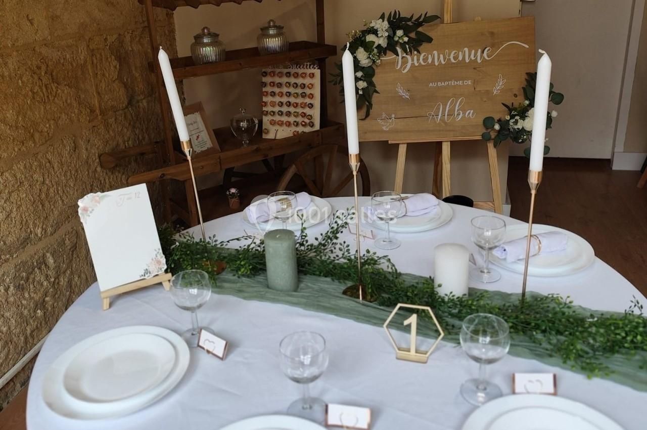 Table décorée pour un baptême avec bougies, vaisselle blanche, chemin de table vert et panneau de bienvenue en arrière-plan.