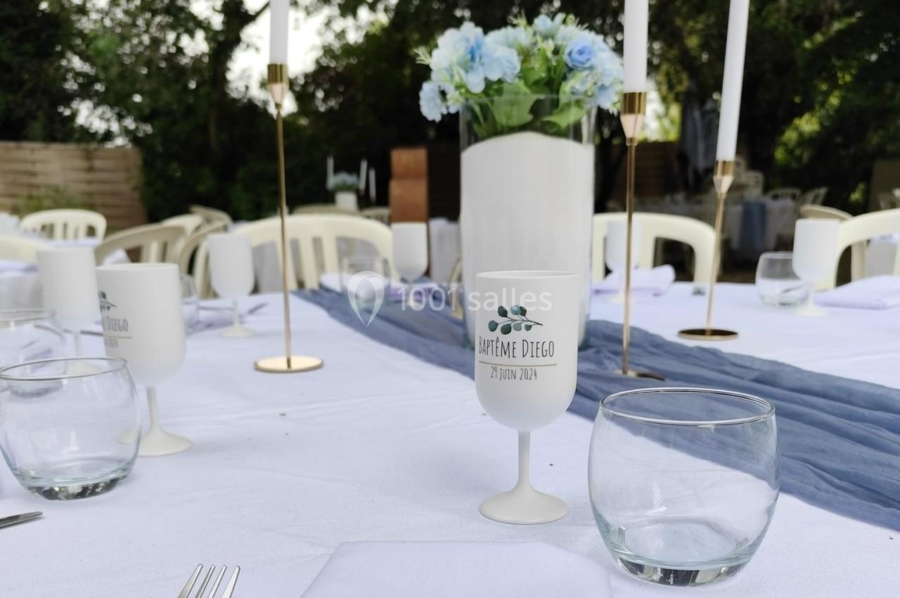 Table dressée pour une réception en extérieur avec nappes blanches, verres, bougies et un vase de fleurs bleues.
