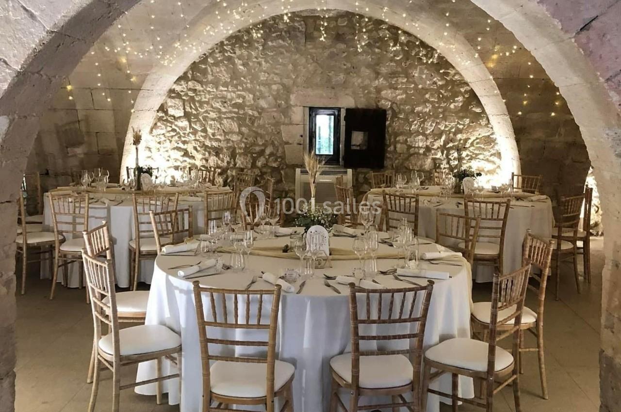 Salle voûtée en pierre décorée pour un événement, avec tables rondes dressées et guirlandes lumineuses suspendues.
