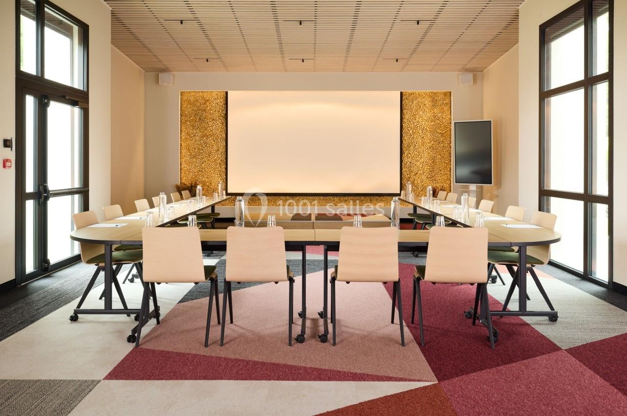 Salle de réunion moderne avec table en U, chaises en bois clair, écran de projection et éclairage chaleureux.