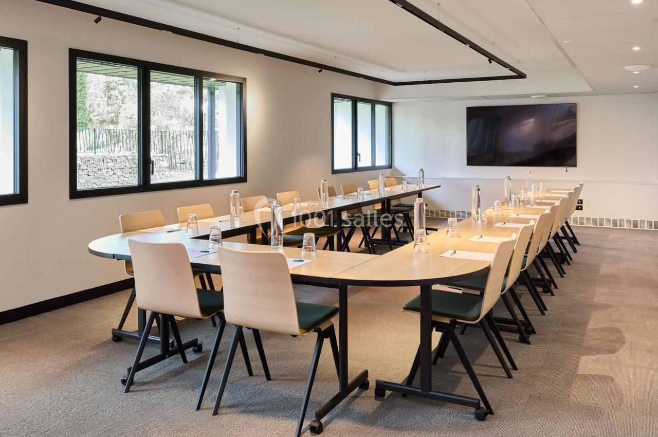Salle de réunion lumineuse avec une grande table en U, des chaises en bois clair et un écran mural.
