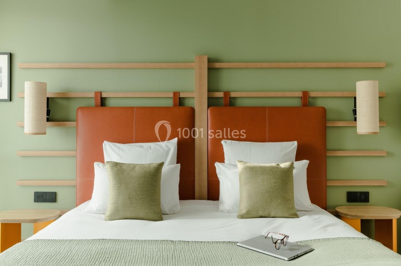 Lit double avec tête de lit en bois, coussins verts, lampe murale et lunettes posées sur une couverture.