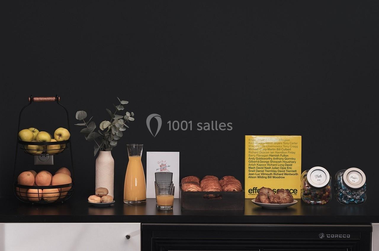 Un comptoir avec des fruits, du jus d'orange, des viennoiseries et des bocaux de friandises devant un mur noir.