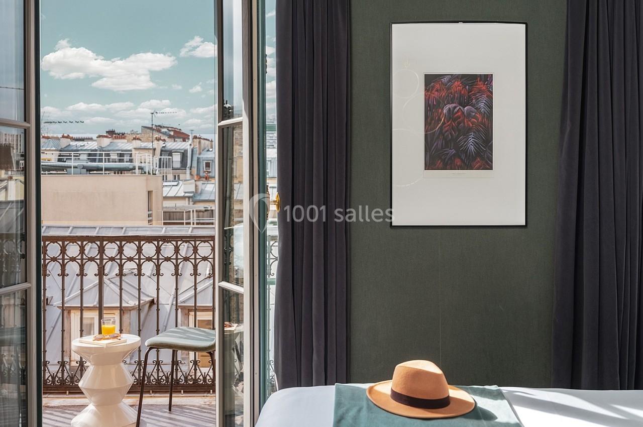 Vue d'une chambre avec balcon, table et chaise, chapeau posé sur le lit, ciel bleu en arrière-plan.