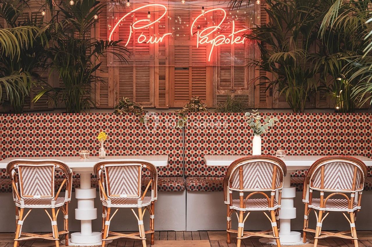 Salle de restaurant avec tables, chaises en rotin, plantes vertes et enseigne néon ’Pour Papoter’ sur un mur décoratif.