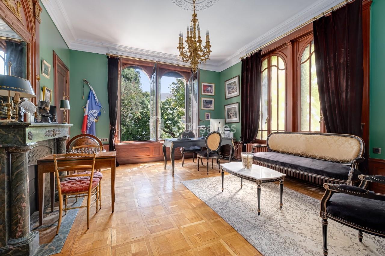 Salon élégant avec parquet, murs verts, grandes fenêtres, mobilier classique et lustre doré, donnant sur un jardin.