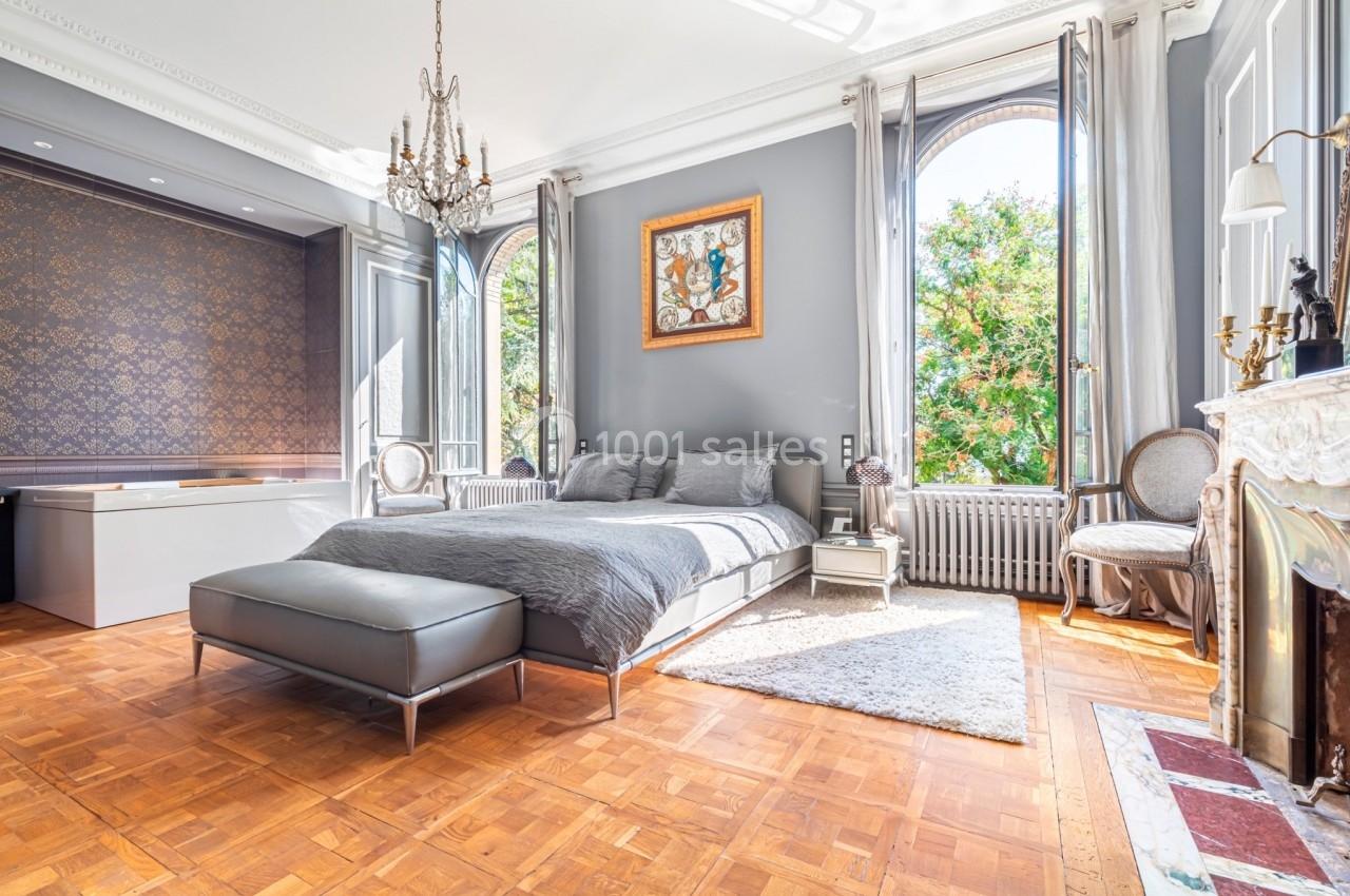 Chambre lumineuse avec lit double, parquet, cheminée, lustre et grandes fenêtres donnant sur des arbres.