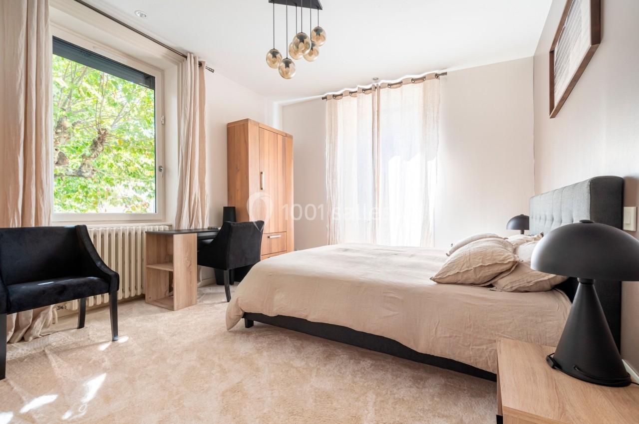 Chambre lumineuse avec lit double, bureau en bois, chaises noires, grandes fenêtres et rideaux beiges.