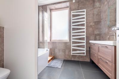 Salle de bain moderne avec carrelage marron, baignoire, fenêtre lumineuse, sèche-serviettes blanc et meuble en bois.
