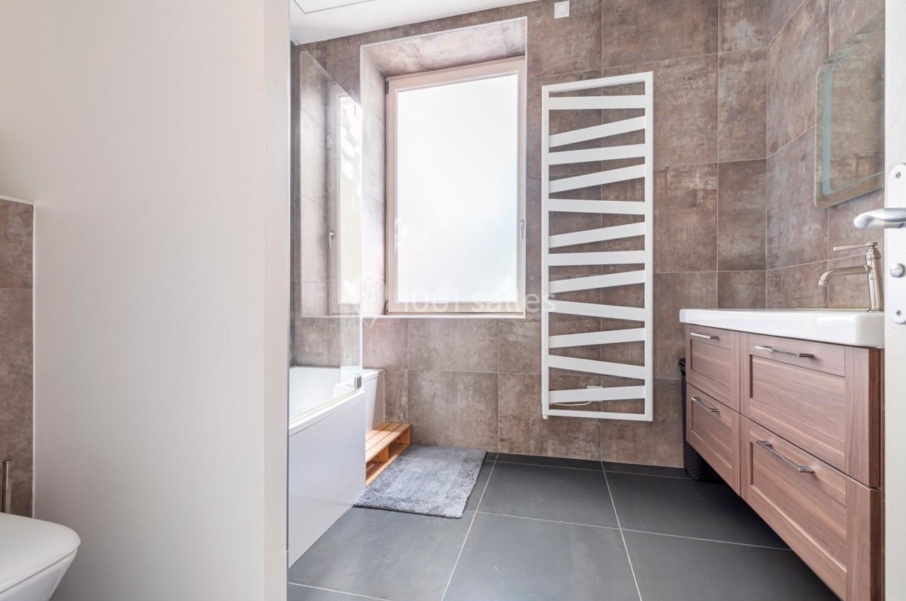 Salle de bain moderne avec carrelage marron, baignoire, fenêtre lumineuse, sèche-serviettes blanc et meuble en bois.