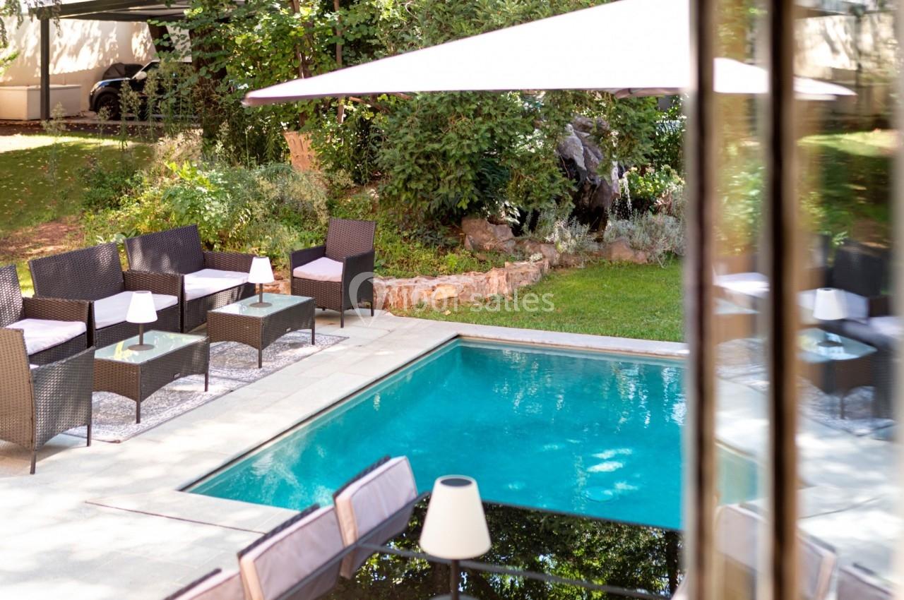 Piscine extérieure entourée de mobilier en rotin, parasol et végétation dans un jardin ensoleillé.
