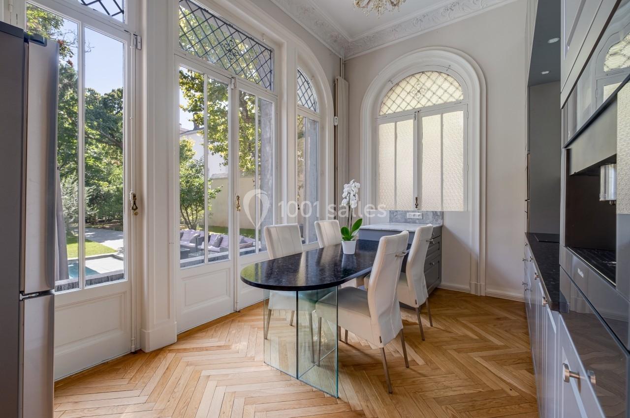 Salle à manger lumineuse avec table en verre, chaises blanches, parquet en bois et grandes fenêtres donnant sur un jardin.