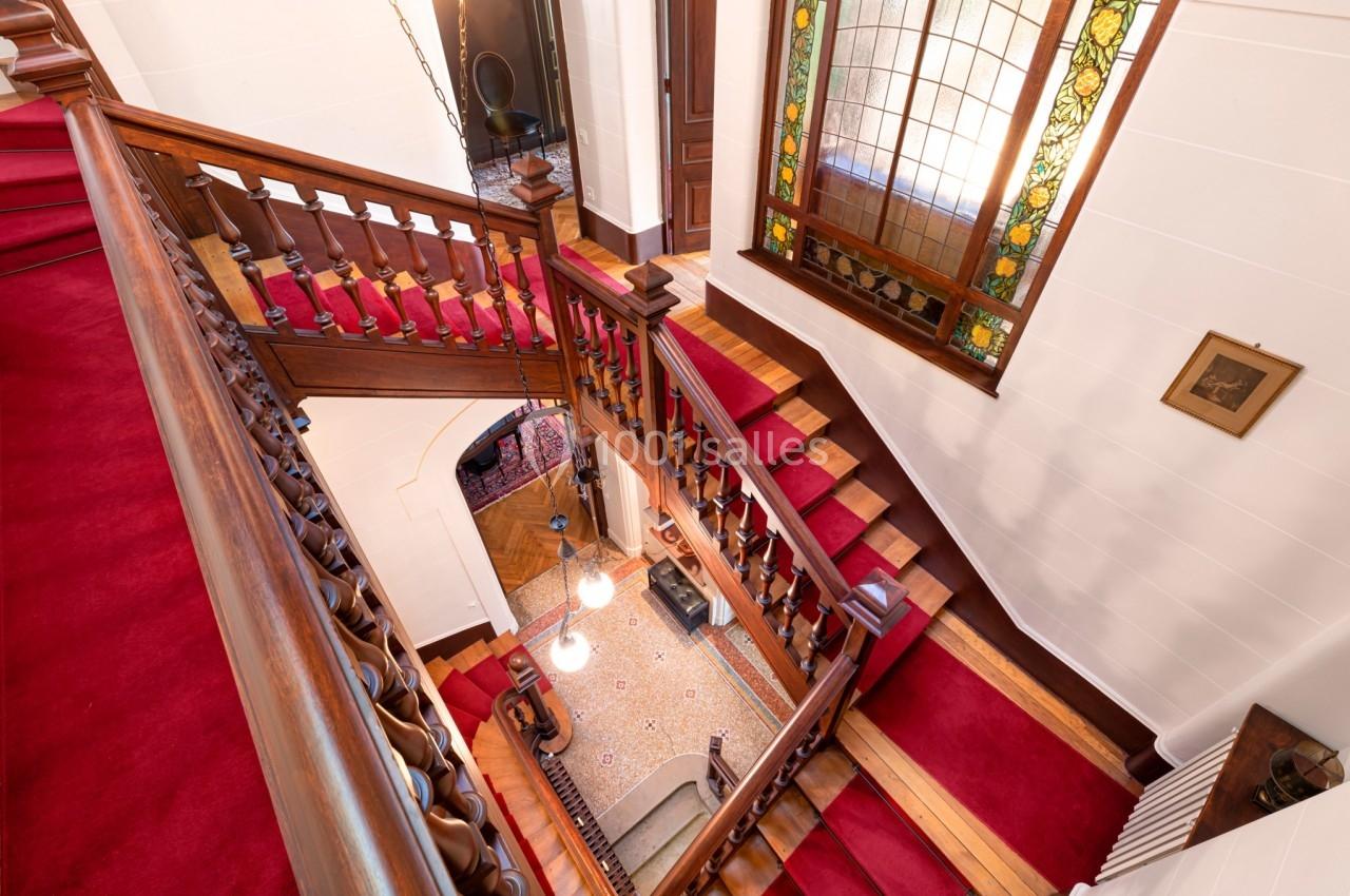 Escalier en bois avec tapis rouge, vitrail coloré et vue plongeante sur un hall éclairé par des suspensions.