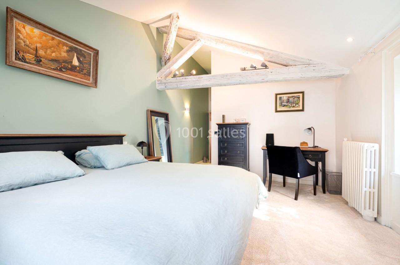 Chambre lumineuse avec lit double, bureau, poutres apparentes et murs verts, décorée avec des tableaux.