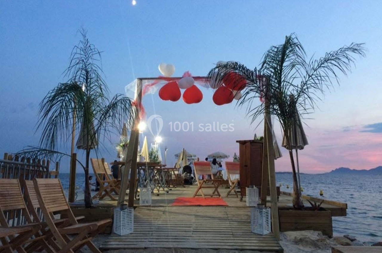 Terrasse en bois décorée avec des chaises, des palmiers et des lampions, donnant sur une plage au coucher du soleil.