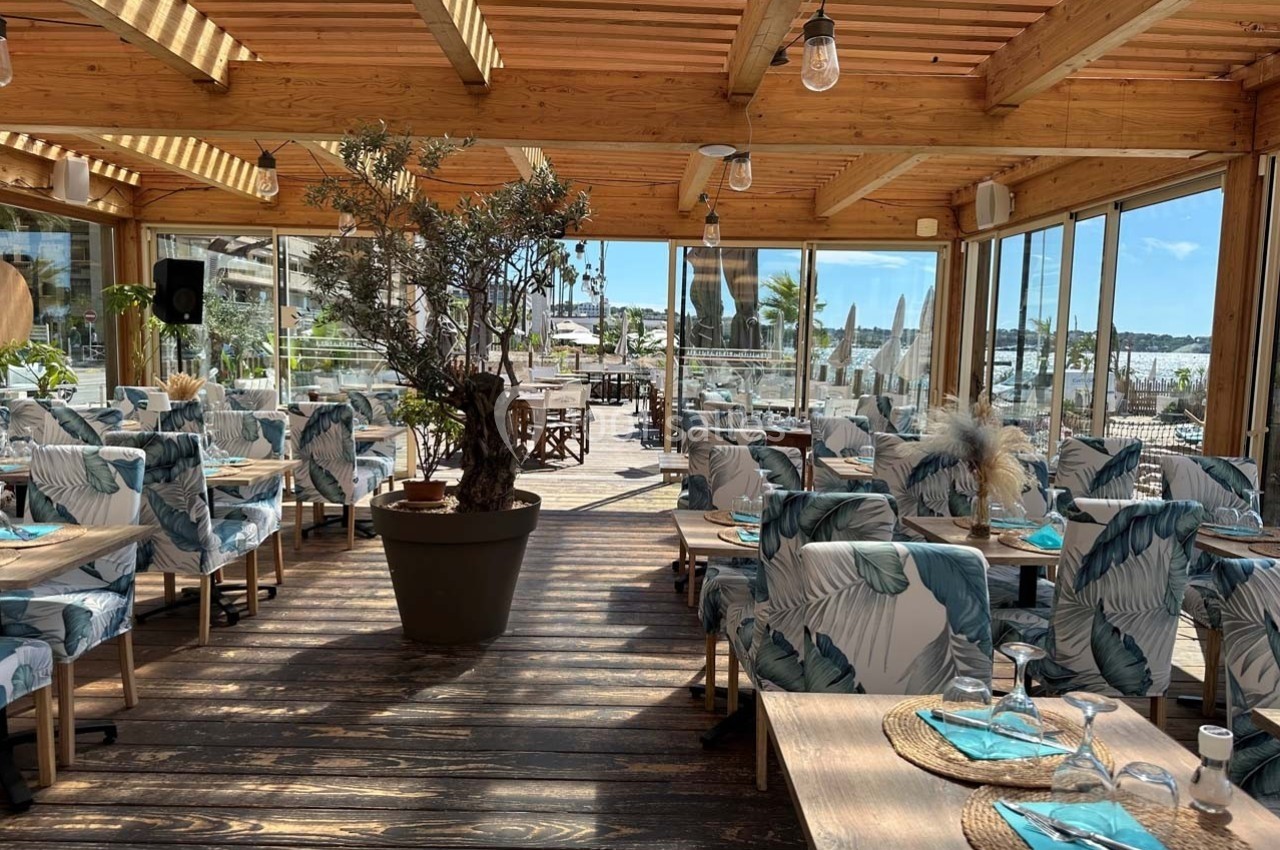 Salle de restaurant lumineuse avec mobilier en bois, chaises à motifs tropicaux et vue sur un port en arrière-plan.