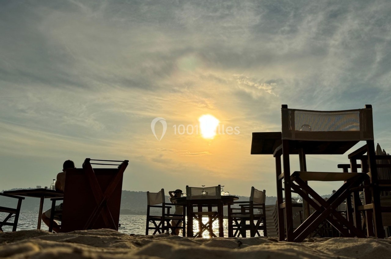 Chaises et tables en bois disposées sur une plage au coucher du soleil, avec vue sur la mer et un ciel partiellement nuageux.