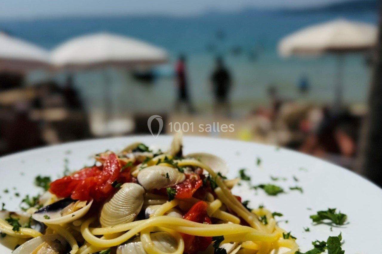 Assiette de pâtes aux palourdes et tomates, garnie de persil, avec une plage floue en arrière-plan.