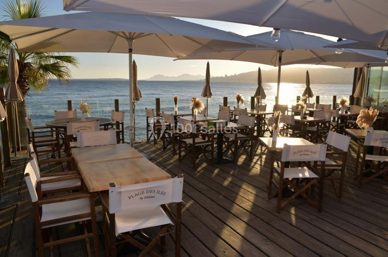 Terrasse en bois d'un restaurant en bord de mer, avec des tables et chaises blanches sous des parasols au coucher du soleil.