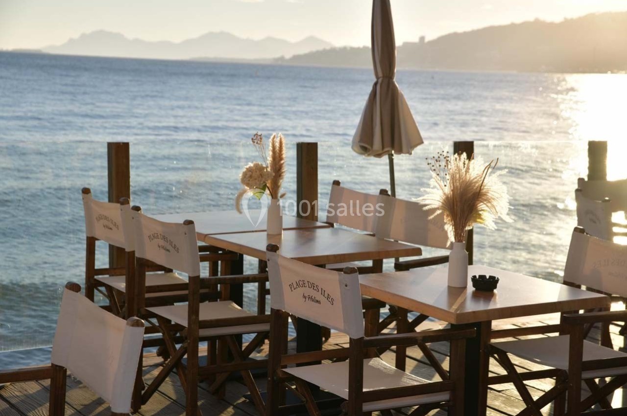 Tables en bois avec chaises pliantes face à la mer, décorées de fleurs séchées, sous une lumière de fin de journée.