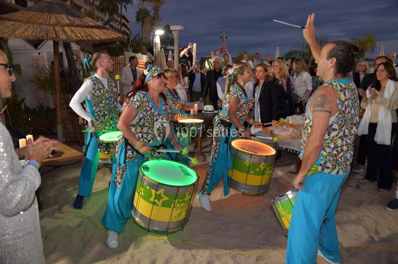 Des musiciens jouent des percussions colorées sur une plage devant un public lors d'un événement en soirée.