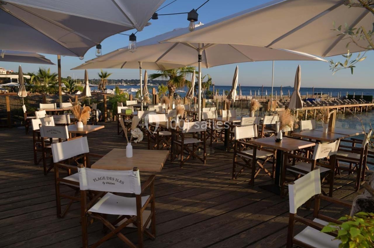 Terrasse en bois avec tables et chaises sous des parasols, offrant une vue sur la mer et un ciel dégagé.