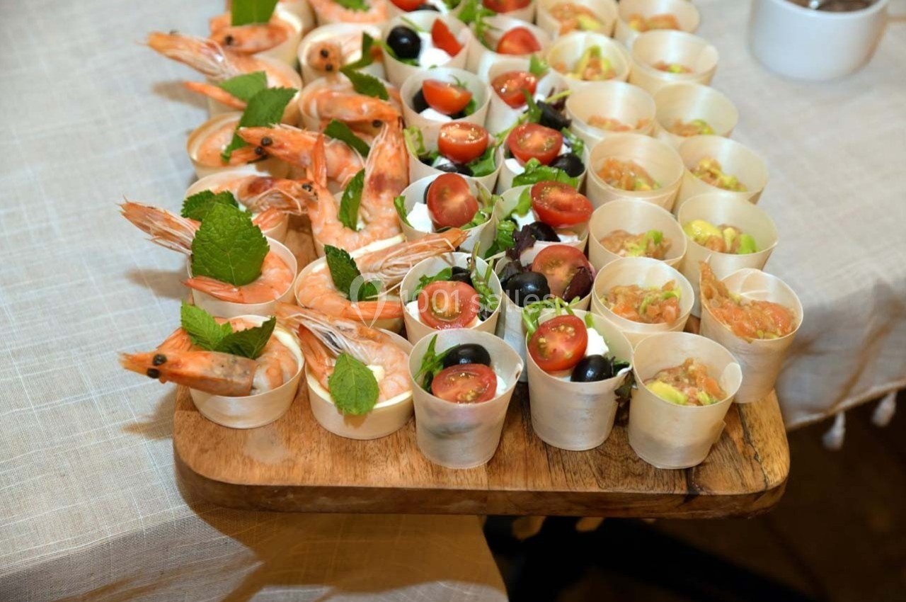 Plateau en bois avec verrines garnies de crevettes, tomates, olives et herbes fraîches, disposées sur une nappe beige.