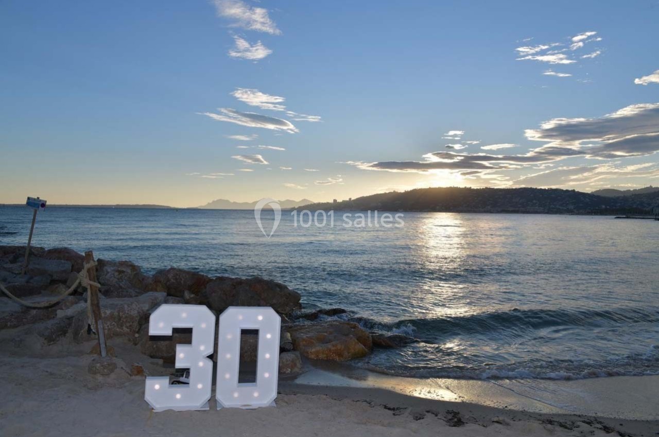 Chiffres lumineux ’30’ posés sur une plage au coucher du soleil, avec vue sur la mer et des collines à l'horizon.