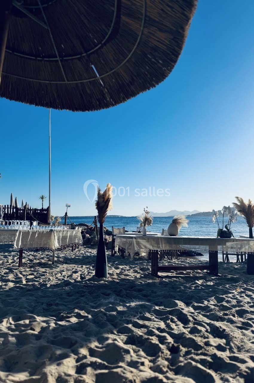 Plage ensoleillée avec tables décorées et parasol en paille, vue sur la mer et les montagnes à l'horizon.