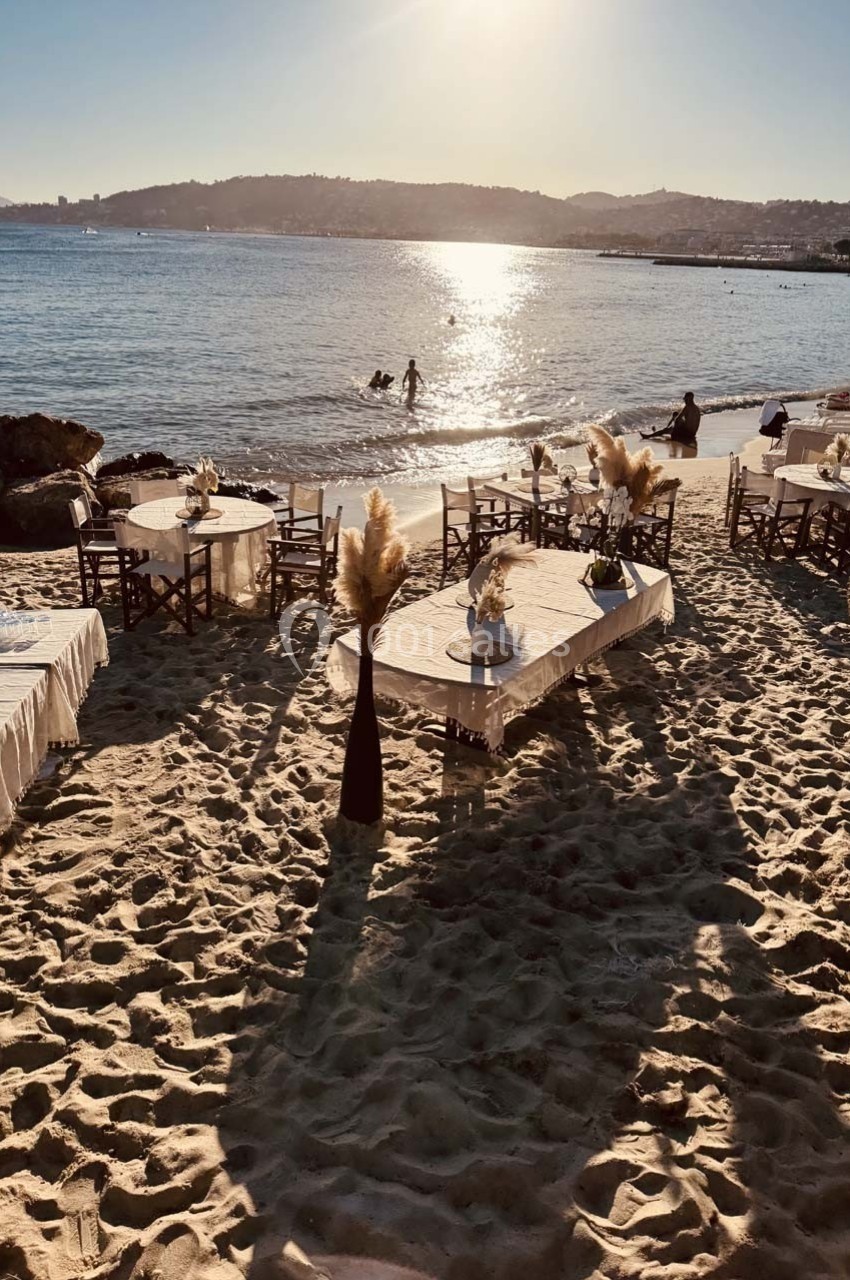 Tables dressées sur une plage de sable au coucher du soleil, avec vue sur la mer et des baigneurs au loin.