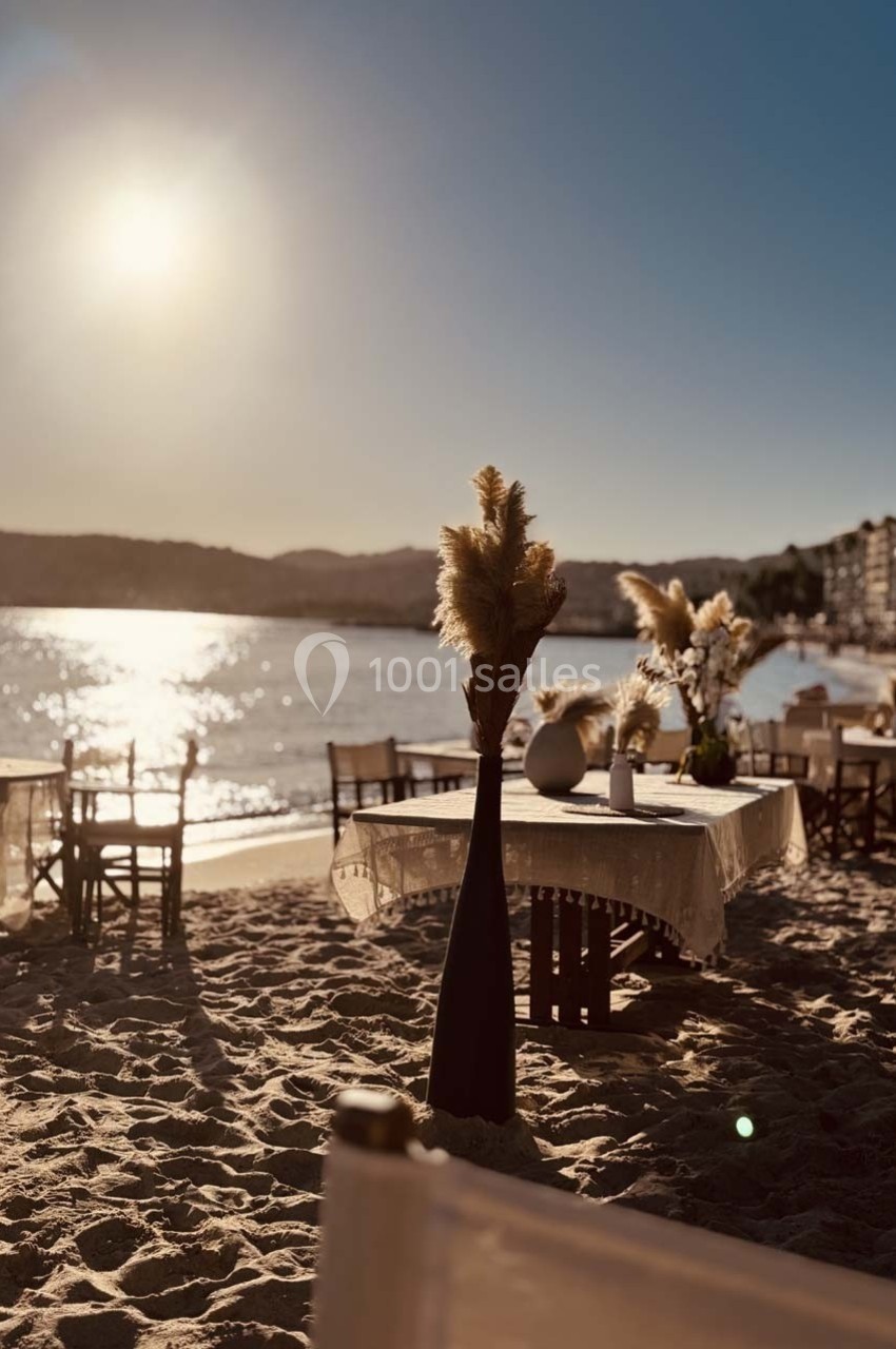 Tables dressées sur une plage au coucher du soleil, avec des décorations florales et une vue sur la mer calme.
