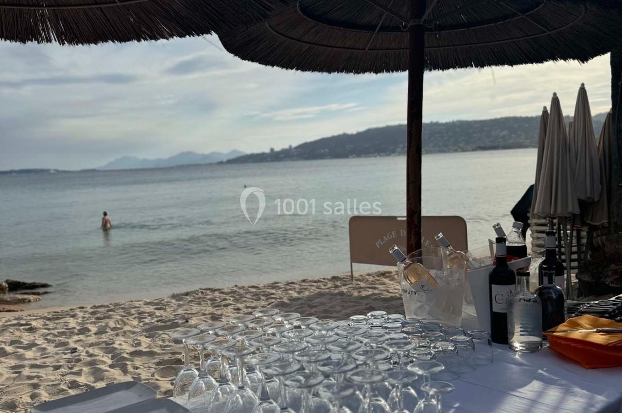 Table dressée avec des verres et des bouteilles sous un parasol en bord de mer, avec une vue sur l'eau et les collines.