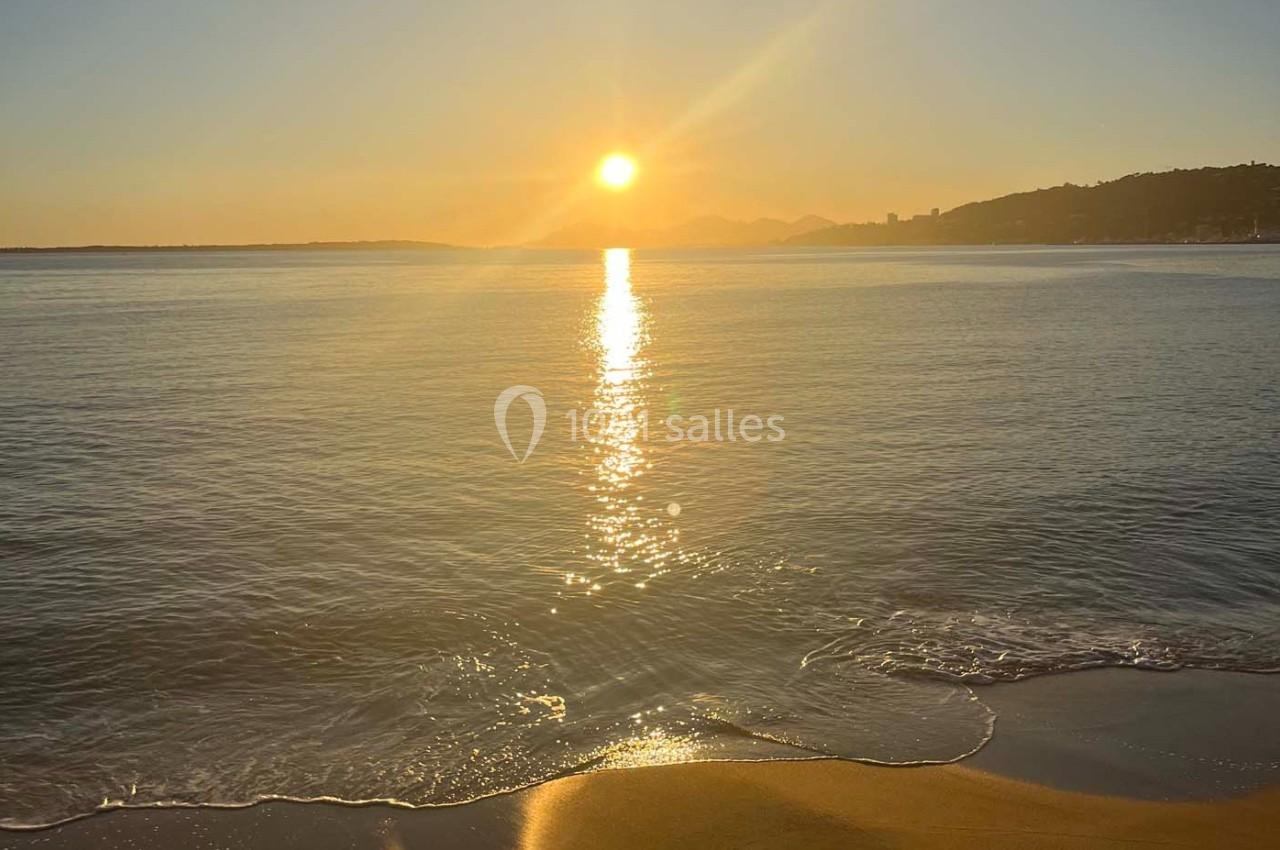 Coucher de soleil sur une plage, avec reflets dorés sur l'eau calme et collines en arrière-plan.
